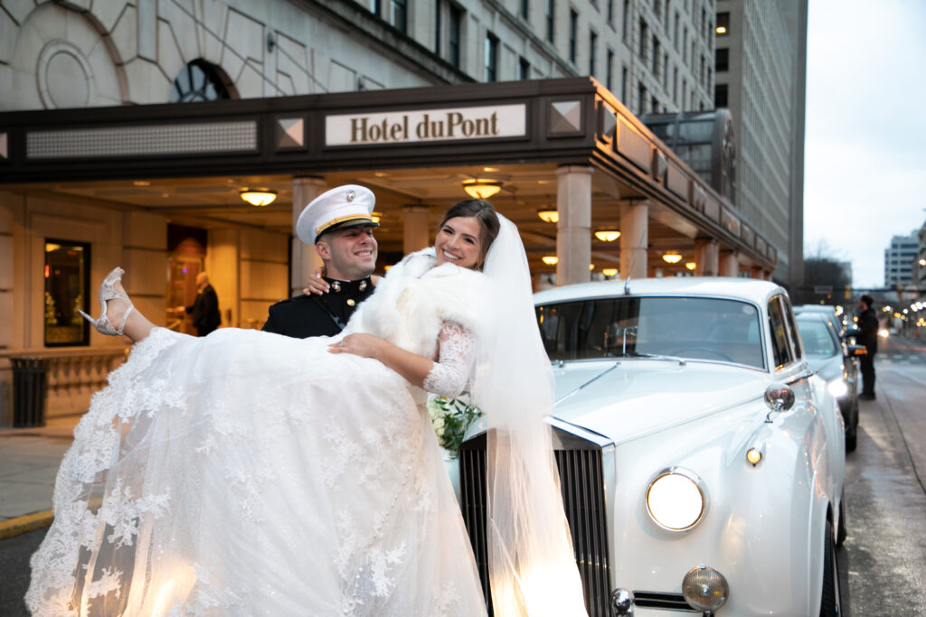 Groom in military dress blues holding a bride in front of a Rolls Royce outside of the Hotel DuPont